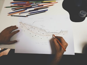 A person is drawing a detailed geological or topographical sketch on white paper. Various colored pencils are scattered above the paper on a white desk. The sketch appears to depict a cross-section of land with annotations.