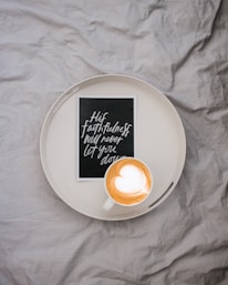 A white circular tray is placed on a crumpled gray fabric surface. Inside the tray are a cup of cappuccino with heart-shaped latte art and a small black card featuring white cursive text that says 'His faithfulness will never let you down.'