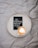 A white circular tray is placed on a crumpled gray fabric surface. Inside the tray are a cup of cappuccino with heart-shaped latte art and a small black card featuring white cursive text that says 'His faithfulness will never let you down.'