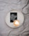 A white circular tray is placed on a crumpled gray fabric surface. Inside the tray are a cup of cappuccino with heart-shaped latte art and a small black card featuring white cursive text that says 'His faithfulness will never let you down.'