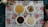 A meal is arranged on a floral-patterned tablecloth with four white plates filled with various foods. The top left plate has a round bread or pancake-type food. The top right plate holds a fresh salad with sliced cucumbers and cherry tomatoes. The bottom left plate contains a dark stew-like dish, possibly with meat or leafy greens. The bottom right plate features a serving of cooked white rice. There are some bottles of beverages or sauces and a couple of drinking glasses also present.