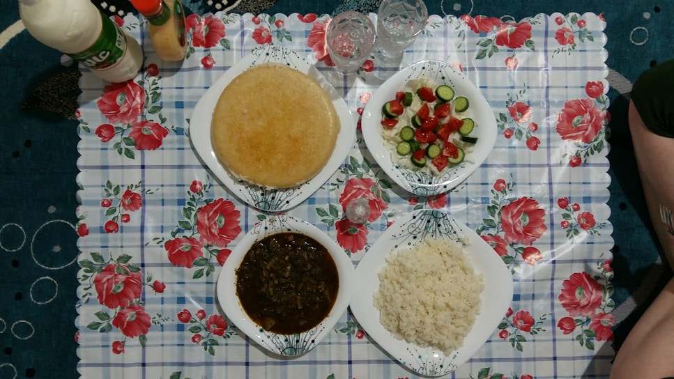 A meal is arranged on a floral-patterned tablecloth with four white plates filled with various foods. The top left plate has a round bread or pancake-type food. The top right plate holds a fresh salad with sliced cucumbers and cherry tomatoes. The bottom left plate contains a dark stew-like dish, possibly with meat or leafy greens. The bottom right plate features a serving of cooked white rice. There are some bottles of beverages or sauces and a couple of drinking glasses also present.
