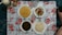A meal is arranged on a floral-patterned tablecloth with four white plates filled with various foods. The top left plate has a round bread or pancake-type food. The top right plate holds a fresh salad with sliced cucumbers and cherry tomatoes. The bottom left plate contains a dark stew-like dish, possibly with meat or leafy greens. The bottom right plate features a serving of cooked white rice. There are some bottles of beverages or sauces and a couple of drinking glasses also present.