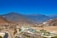 A picturesque landscape featuring an airport nestled in a valley surrounded by mountains. The clear blue sky contrasts with the earthy tones of the hills and the modern architecture of the airport buildings. A river winds along a road in the foreground, with some residential structures scattered throughout the valley.