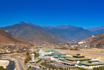 A picturesque landscape featuring an airport nestled in a valley surrounded by mountains. The clear blue sky contrasts with the earthy tones of the hills and the modern architecture of the airport buildings. A river winds along a road in the foreground, with some residential structures scattered throughout the valley.