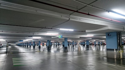 An underground parking garage with multiple parking spaces lined by blue columns. The area is well-lit with fluorescent lights, reflecting off the polished concrete floor. Cars are parked in some spaces while others remain empty. Signs indicate exit directions and specific sections of the garage.