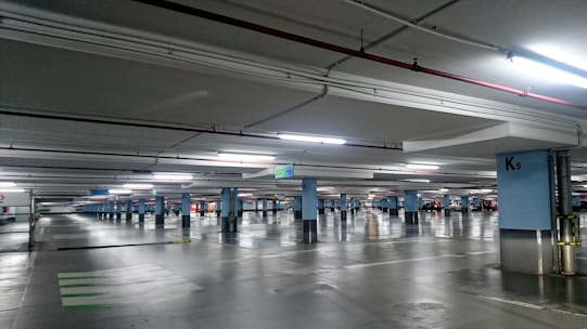 An underground parking garage with multiple parking spaces lined by blue columns. The area is well-lit with fluorescent lights, reflecting off the polished concrete floor. Cars are parked in some spaces while others remain empty. Signs indicate exit directions and specific sections of the garage.