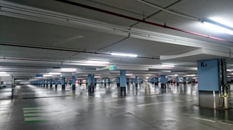 An underground parking garage with multiple parking spaces lined by blue columns. The area is well-lit with fluorescent lights, reflecting off the polished concrete floor. Cars are parked in some spaces while others remain empty. Signs indicate exit directions and specific sections of the garage.
