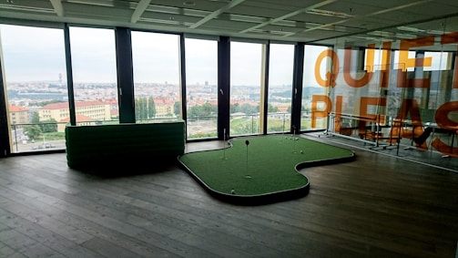A modern office room with large floor-to-ceiling windows offering a panoramic city view. The room features a mini-golf putting green with golf balls. There is a glass wall with the words 'QUIET PLEASE' in large orange letters. The floor is dark wood, and there is a green cushioned seating area near the windows.