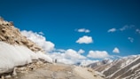 A rocky mountain road with patches of snow on the sides and a motorbike traveling along it. The sky is clear and blue with a few fluffy clouds scattered throughout. Snow-capped mountains are visible in the background, emphasizing the high-altitude setting.