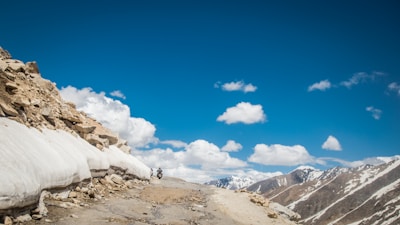 A rocky mountain road with patches of snow on the sides and a motorbike traveling along it. The sky is clear and blue with a few fluffy clouds scattered throughout. Snow-capped mountains are visible in the background, emphasizing the high-altitude setting.