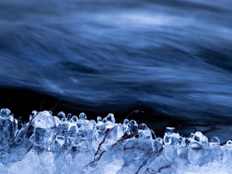 Clear, glossy ice formations are captured in the foreground, with delicate, unique shapes that appear almost sculpted. The background consists of a smooth, flowing stream of water, creating a striking contrast between the chaotic, frozen foreground and the calm, fluid flow of water.