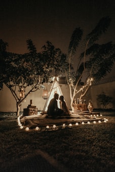 A cozy nighttime outdoor scene featuring a couple sitting inside a small illuminated tent under a tree. String lights are gently illuminating the surrounding area, enhancing the romantic atmosphere. Various decorative items and bottles are placed around the tent, with trees and grass visible in the background.
