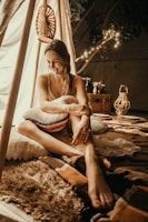 A woman is sitting inside a cozy, warmly lit tent. She is smiling and appears relaxed, surrounded by soft pillows and rugs. Decorative lanterns and string lights add to the ambient mood. The setting suggests an outdoor or backyard camping experience.