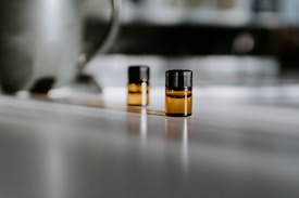 Two small amber glass vials with black caps are placed on a smooth surface, with soft lighting creating shadows and reflections. The background is blurred, emphasizing the vials.
