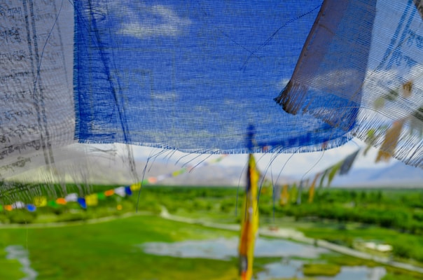 Colorful Tibetan prayer flags gently flutter, creating a vivid tapestry against a backdrop of a lush, green landscape and blue sky. The fabric is semi-transparent, allowing glimpses of distant mountains and a serene field with patches of water. The arrangement suggests a sacred or tranquil location, enlivened by the lively dance of the flags in the breeze.