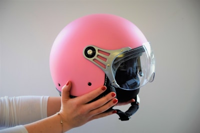 A person holds a pink motorcycle helmet with a clear visor. The helmet features metallic detailing on the side. The individual wears a white long-sleeved shirt, and their nails are painted red.