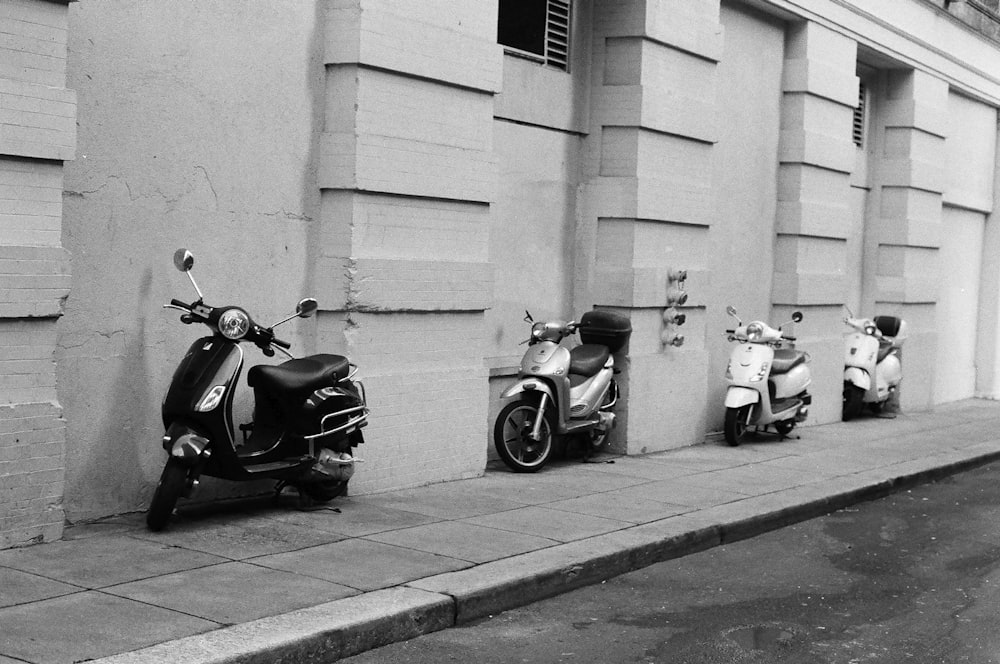 grayscale photography of four motor scooter park beside concrete wall