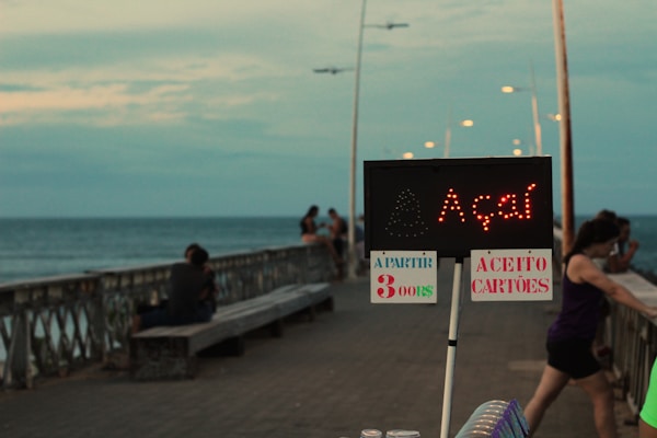 A wooden pier extends out over the ocean, with people leisurely sitting and standing along the railing. A digital sign with red letters displays the word açaí, accompanied by handwritten signs indicating prices and payment methods. Streetlights line the pier, leading toward the horizon on a cloudy day.