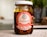 A glass jar filled with salsa de chile de árbol with a white lid. The label is prominently displayed in red and white, featuring the brand name 'alpizar' and the weight of 235g. The ingredients inside the jar are visible through the glass, showcasing a chunky, dark red salsa with chili pieces. The background is softly blurred, suggesting an indoor setting with some greenery.
