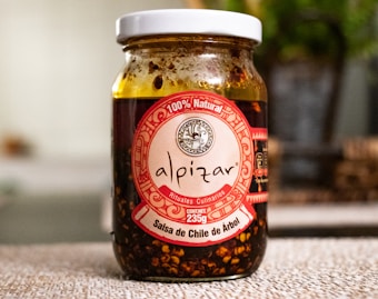 A glass jar filled with salsa de chile de &aacute;rbol with a white lid. The label is prominently displayed in red and white, featuring the brand name 'alpizar' and the weight of 235g. The ingredients inside the jar are visible through the glass, showcasing a chunky, dark red salsa with chili pieces. The background is softly blurred, suggesting an indoor setting with some greenery.