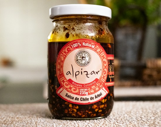 A glass jar filled with salsa de chile de &aacute;rbol with a white lid. The label is prominently displayed in red and white, featuring the brand name 'alpizar' and the weight of 235g. The ingredients inside the jar are visible through the glass, showcasing a chunky, dark red salsa with chili pieces. The background is softly blurred, suggesting an indoor setting with some greenery.