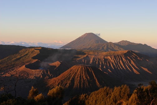 A stunning landscape of volcanic mountains at sunrise, with clear skies and gentle sunlight casting long shadows over the rugged terrain. Sparse vegetation covers some parts of the slopes, and a light mist or smoke rises from the caldera, adding an ethereal quality to the scene.