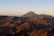 A stunning landscape of volcanic mountains at sunrise, with clear skies and gentle sunlight casting long shadows over the rugged terrain. Sparse vegetation covers some parts of the slopes, and a light mist or smoke rises from the caldera, adding an ethereal quality to the scene.