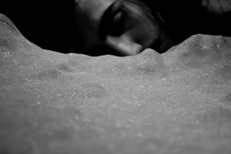 A close-up black and white image focusing on a textured surface in the foreground. In the background, there is a partially visible face of a person, appearing to be sleeping or resting.