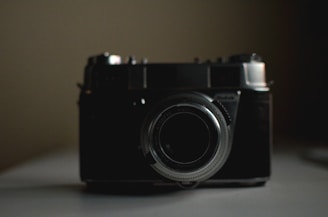 A vintage film camera is positioned centrally with a slightly dark and subdued background. The camera is highlighted with a soft light that accentuates its metallic lens and body details.
