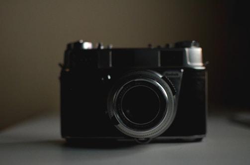 A vintage film camera is positioned centrally with a slightly dark and subdued background. The camera is highlighted with a soft light that accentuates its metallic lens and body details.