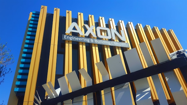 A modern building with a prominent sign that reads 'AXON Engineering & Building Company'. The structure features vertical yellow and white beams against a deep blue sky.