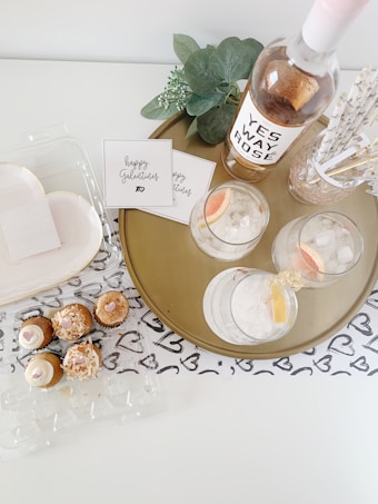A round golden tray holds a bottle of rose wine labeled 'YES WAY ROSE', two glasses filled with ice and grapefruit slices, decorative straws in a container, and a small bouquet of eucalyptus leaves. Beside the tray, there are cards with 'Happy Galentine's' written on them, a light-colored surface with a heart-patterned cloth underneath, and a clear plastic container with assorted cupcakes, each topped with icing and toppings.