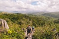 Several people are hiking along a narrow, rocky path surrounded by dense green vegetation. The landscape extends into rolling hills covered with lush forests under a partly cloudy sky. The path appears to be at an elevated position offering a panoramic view of the expansive landscape.
