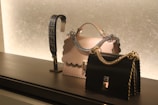 On a sleek black surface, a display features two elegantly designed handbags. The central bag is cream-colored with a scalloped edge and a silver chain handle, while the other is black with a gold chain. To the left, there's a decorative strap showcasing an intricate design.