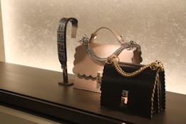 On a sleek black surface, a display features two elegantly designed handbags. The central bag is cream-colored with a scalloped edge and a silver chain handle, while the other is black with a gold chain. To the left, there's a decorative strap showcasing an intricate design.