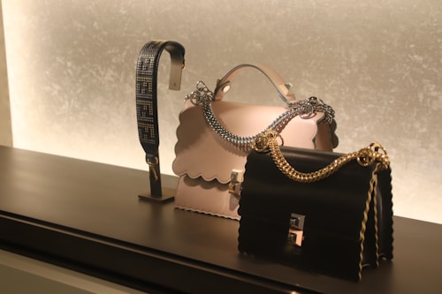On a sleek black surface, a display features two elegantly designed handbags. The central bag is cream-colored with a scalloped edge and a silver chain handle, while the other is black with a gold chain. To the left, there's a decorative strap showcasing an intricate design.