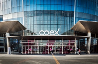 The image features a modern building facade with a large glass entrance and the word 'coex' prominently displayed above the doors. The structure has a sleek and urban design with metal and glass elements. People are walking in front of the building, emphasizing its function as a public or commercial space.