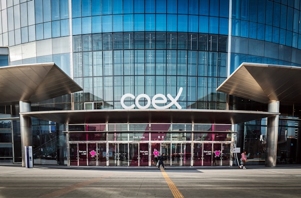 The image features a modern building facade with a large glass entrance and the word 'coex' prominently displayed above the doors. The structure has a sleek and urban design with metal and glass elements. People are walking in front of the building, emphasizing its function as a public or commercial space.