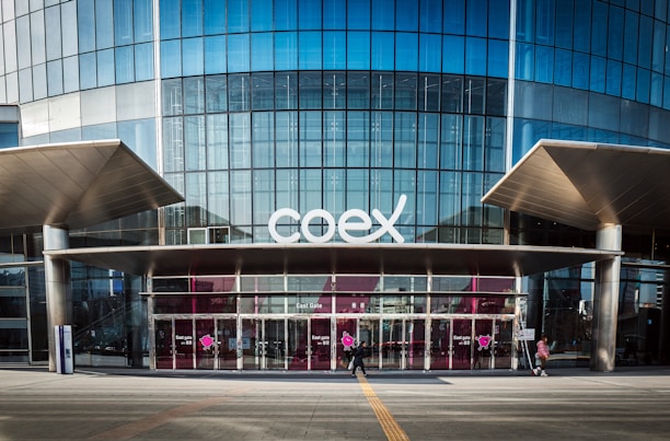The image features a modern building facade with a large glass entrance and the word 'coex' prominently displayed above the doors. The structure has a sleek and urban design with metal and glass elements. People are walking in front of the building, emphasizing its function as a public or commercial space.