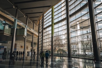 A spacious modern building with large glass windows stretching from floor to ceiling, allowing abundant natural light to flood the area. Several people are walking through the polished floors, and the architecture includes metallic pillars and elevated walkways. Outside, trees and other buildings can be seen, contributing to an urban setting.