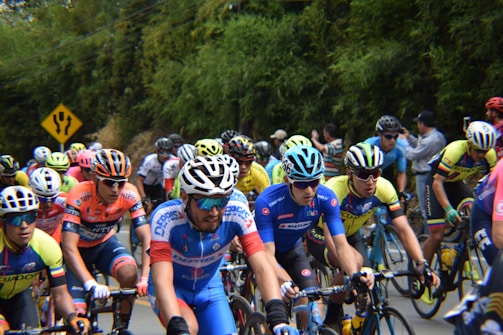 A group of competitive cyclists dressed in brightly colored jerseys and helmets are racing on a road surrounded by greenery. Spectators can be seen in the background. A yellow road sign indicating a fork is visible.