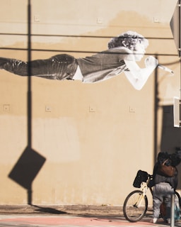 A large mural of a person with curly hair is painted on a beige wall. This person is depicted in a horizontal position, holding a microphone. Below the mural, a person stands next to a beige bicycle, their back turned to the camera. Long shadows and a sense of depth are created by the lighting.