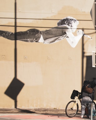 A large mural of a person with curly hair is painted on a beige wall. This person is depicted in a horizontal position, holding a microphone. Below the mural, a person stands next to a beige bicycle, their back turned to the camera. Long shadows and a sense of depth are created by the lighting.