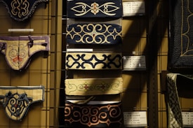 Various intricately designed ornamental textiles displayed on a metal grid, featuring elaborate patterns and vibrant colors. Each piece is tagged with informational labels in what appears to be a museum or gallery setting. The textiles use rich blue, cream, and black colors, showcasing a variety of swirling and symmetrical designs.