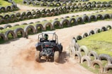 A rugged off-road vehicle navigates a winding dirt track, bordered by rows of repurposed tires serving as barriers. A driver is seen wearing a helmet, maneuvering the vehicle with dirt being kicked up from the wheels. Grass grows between the tire barriers, adding greenery to the scene.