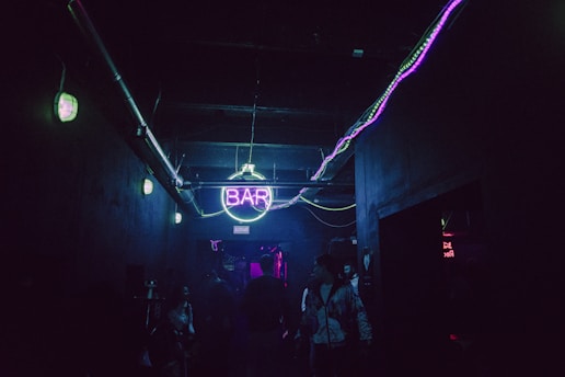 A dimly lit hallway features a neon 'BAR' sign glowing in purple and green. The dark walls and ceiling are accented by horizontal light strips. Several people are gathered, partially visible, creating a lively atmosphere.