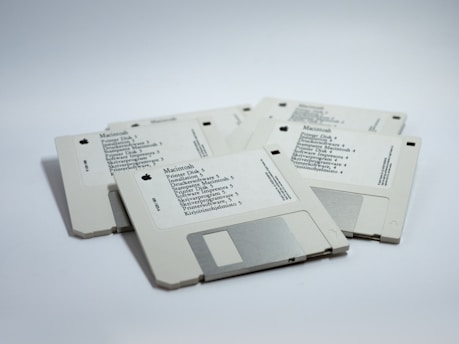 A group of five vintage Macintosh floppy disks arranged in a loose pile on a white surface. Each disk has a label indicating various software titles and installation information. The labels are printed in black text with small Apple logos, and the disks are a light gray color with a metal shutter.