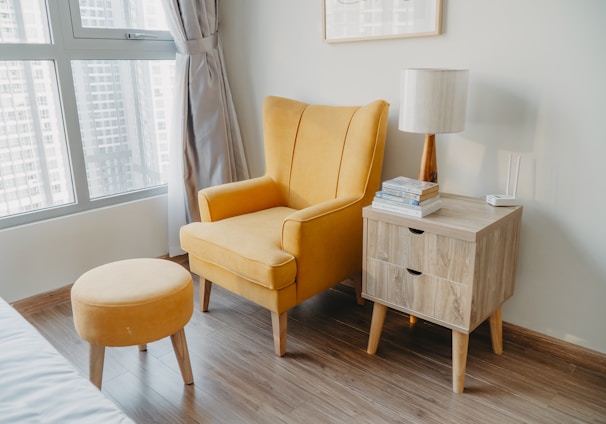 yellow armchair and stool beside wooden nightstand by the wall near glass window and bed