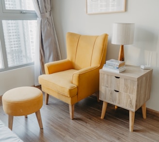 yellow armchair and stool beside wooden nightstand by the wall near glass window and bed